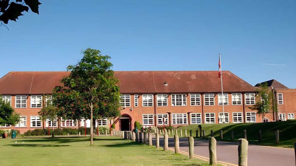 Das mehrstöckige Backsteingebäude mit rotem Dach des Brockenhurst College steht in einer Grünfläche mit Bäumen und blauem Himmel.