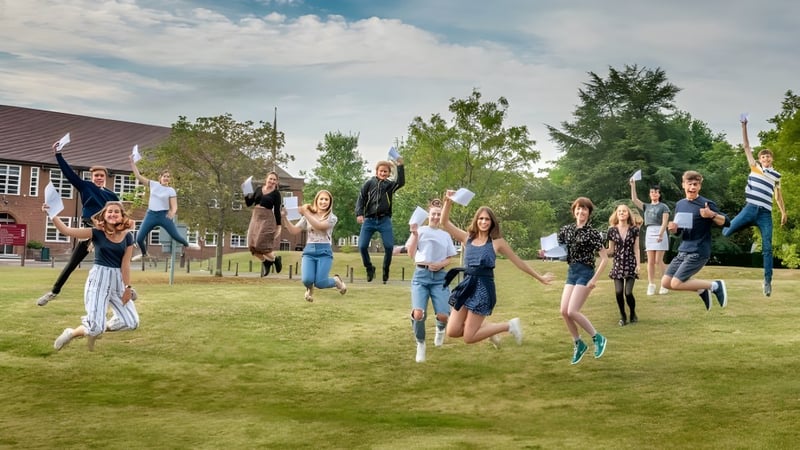 Eine Gruppe Schülerinnen und Schüler des Brockenhurst College springt und feiert auf einer Wiese vor Bäumen und Gebäuden.