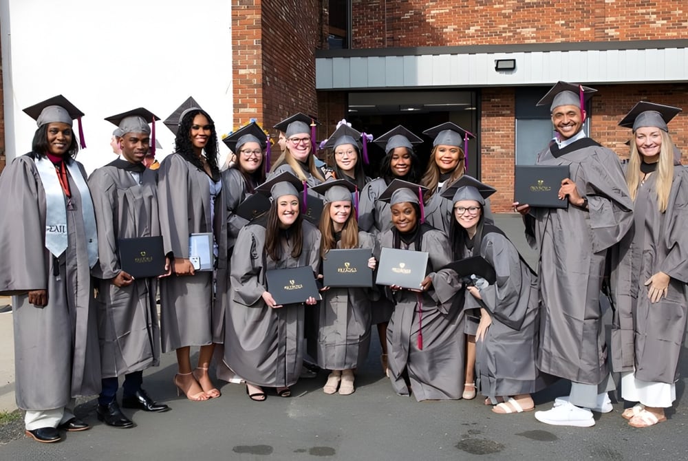 Eine Gruppe Absolventinnen und Absolventen in grauem Gewand steht vor einem Backsteingebäude auf dem Campus des Brookdale Community College und hält ihre Diplome.