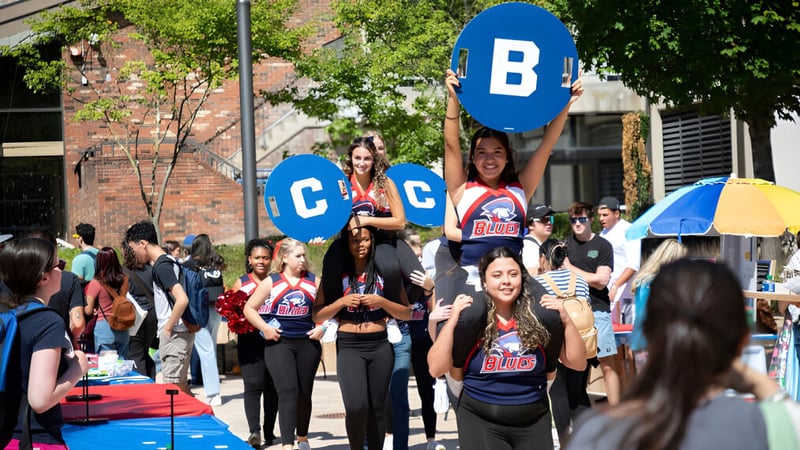 Eine Gruppe von Schülern hält bunte Schilder vor dem Backsteingebäude des Brookdale Community College an einem sonnigen Tag.