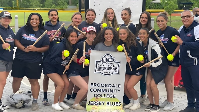 Eine Gruppe junger Studentinnen des Brookdale Community College posiert mit dem Softball-Team-Banner auf einem grasbewachsenen Spielfeld.