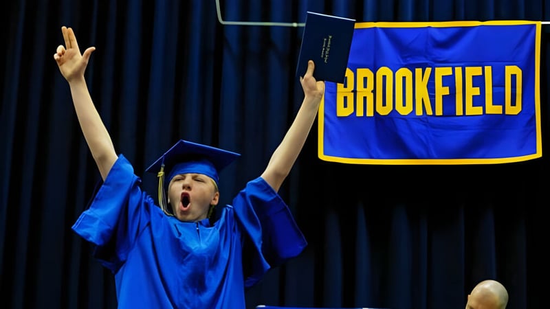 Eine Person in blauer Abschlussrobe hebt vor einem Banner der Brookfield High School die Arme zum Jubel.