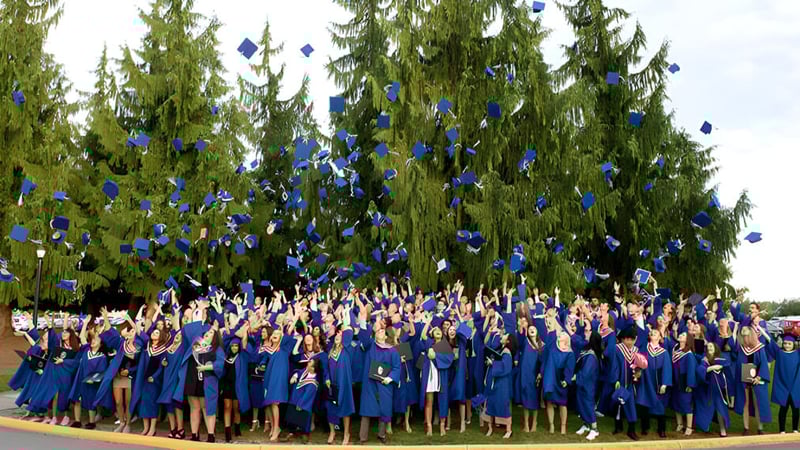 Eine Gruppe von Absolventen in blauen Talaren steht vor hohen immergrünen Bäumen auf dem Gelände der Brookswood Secondary School.