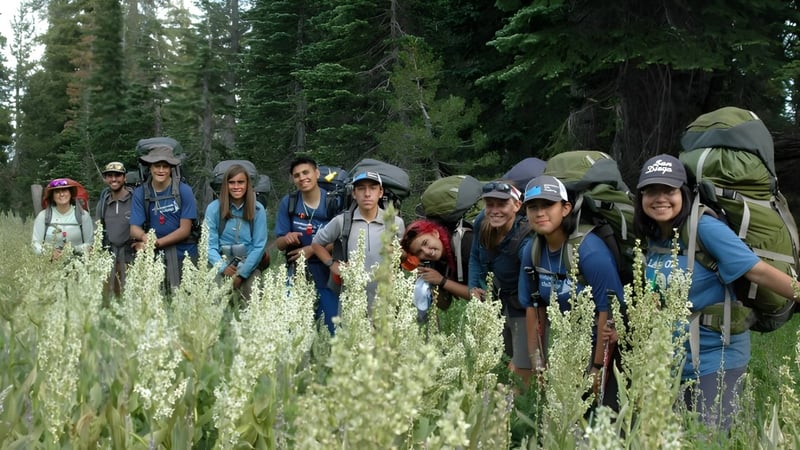 Schüler der Buckswood School stehen mit Wanderausrüstung auf einer Wiese mit hohem Gras vor einem dichten Wald.