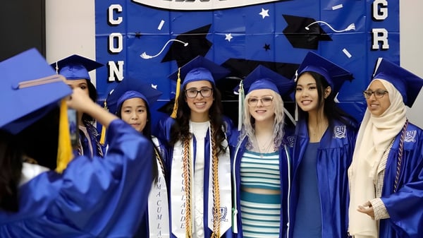 Schülerinnen der Burlington Central High School stehen in Abschlusskleidung vor einer Kulisse mit dem Schriftzug Congratulations.