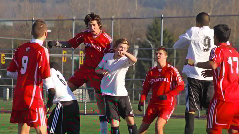 Schüler der Burnaby Central School spielen Fußball auf einem Rasenfeld vor einem Zaun und Bäumen.