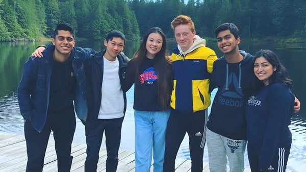 Eine Gruppe Schülerinnen und Schüler der Burnaby Mountain School steht auf einem Holzsteg vor einem ruhigen See mit grünen Bäumen.