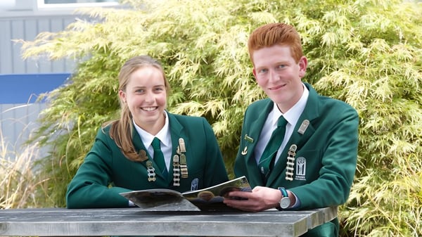 Zwei Schülerinnen und Schüler in grünen Schuluniformen stehen gemeinsam auf dem Gelände der Burnside High School vor grünem Laub.