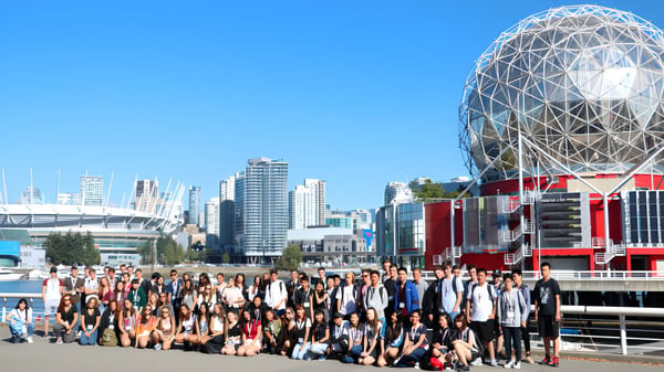 Eine Gruppe von Schülerinnen und Schülern versammelt sich vor dem modernen Kuppelgebäude der Burnsview Secondary School vor der Stadtlandschaft.