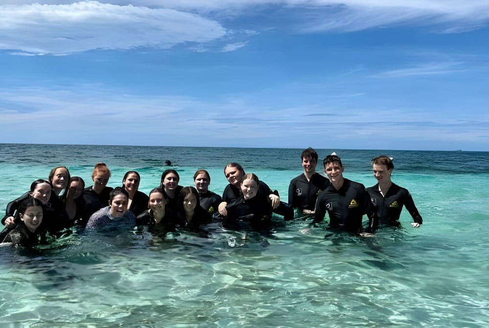 Eine Gruppe von Schülern des Burpengary State Secondary College steht in Neoprenanzügen im flachen, türkisfarbenen Wasser eines tropischen Strandes.