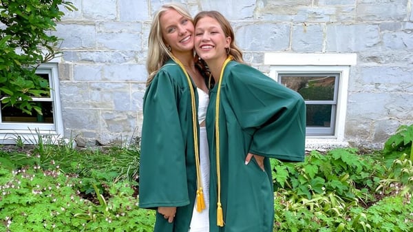 Zwei Schülerinnen der Burr and Burton Academy in grünen Abschlussroben umarmen sich vor einem Steingebäude mit grüner Umgebung.