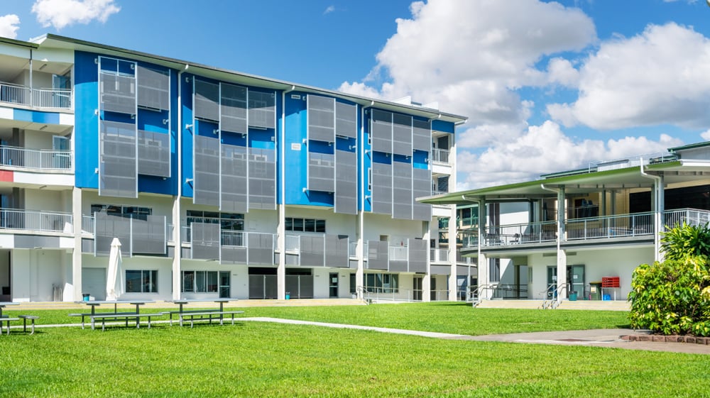 Das moderne mehrstöckige Gebäude mit bunter Glasfassade der Bush Post Primary School steht in einer gepflegten Grünanlage unter blauem Himmel.