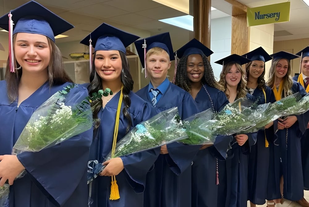 Absolventen der Cadillac Heritage Christian School stehen im Flur mit blauen Abschlussgewändern und Blumensträußen.