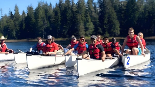 Schüler der Caledonia Secondary School sitzen mit roten Rettungswesten in kleinen weißen Booten auf einem See in einem bewaldeten Gebiet.