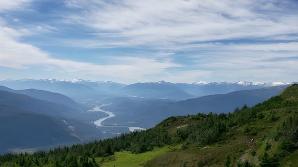 Eine grüne Berglandschaft mit einem Fluss und schneebedeckten Gipfeln im Hintergrund nahe der Caledonia Secondary School.
