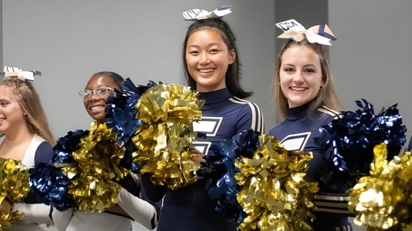 Eine Gruppe Cheerleader in Marine- und Golduniformen mit Pompons posiert gemeinsam auf dem Campus der Calvary Christian Academy.
