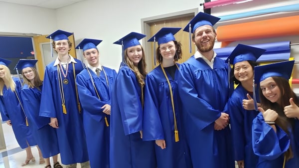 Eine Gruppe von Absolventen der Cambridge Christian School steht im Flur in blauen Abschlusskleidern und -mützen für ein Gruppenfoto.