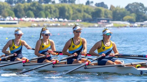 Vier Ruderinnen auf dem Wasser vor einer Kulisse aus Bäumen und Gebäuden auf dem Gelände der Cambridge High School.