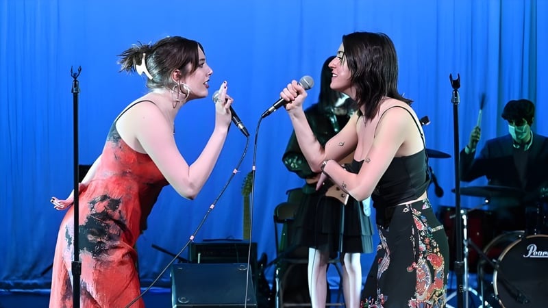 Zwei Schülerinnen führen auf der Bühne der The Cambridge School of Weston vor blauem Hintergrund mit Musikinstrumenten auf.