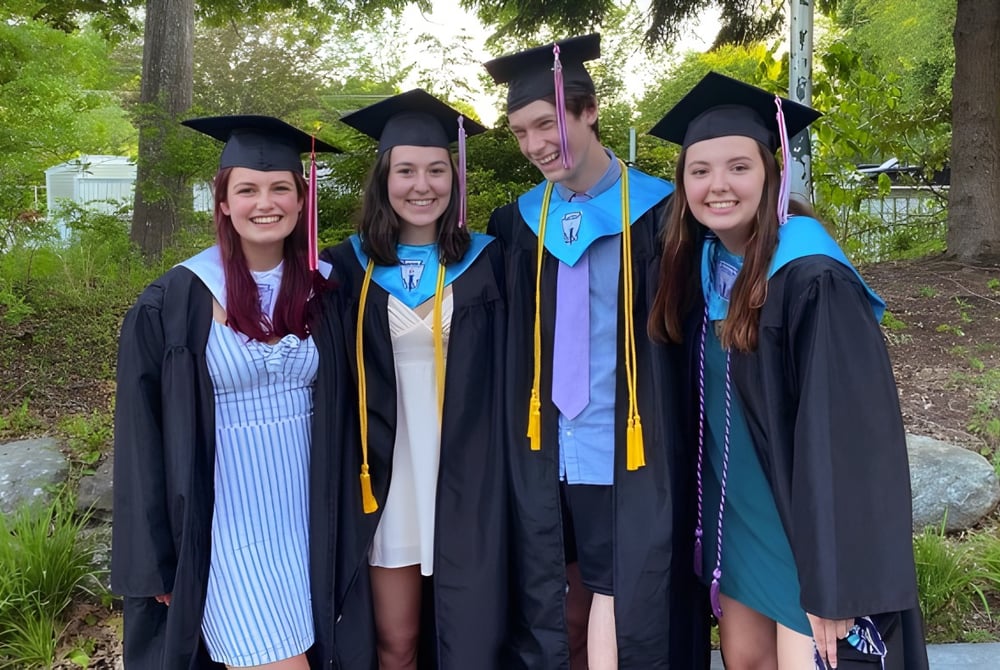 Eine Gruppe von vier jungen Frauen in Abschlusskleidung steht auf dem Campus der Camden Hills Regional High School im Grünen.