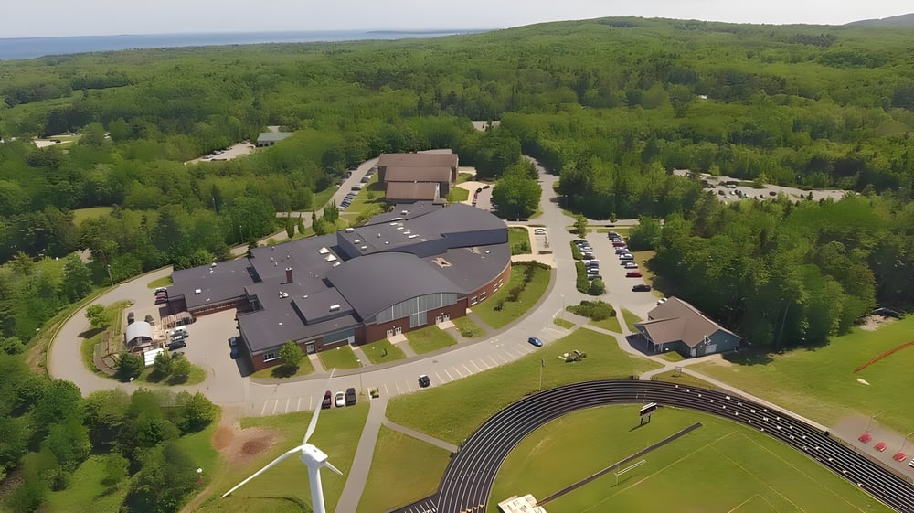 Das mehrstöckige Gebäude der Camden Hills Regional High School steht in einer bewaldeten Landschaft mit einer Zufahrtsstraße und Windrädern im Hintergrund.