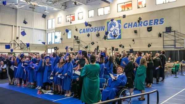 Eine große Gruppe von Absolventen in blauen und grünen Roben versammelt sich in einer Sporthalle auf dem Campus des Camosun College.