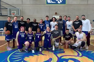 Eine Gruppe von Basketballspielern posiert auf dem Basketballfeld im Camosun College vor einem blau-weißen Logo an der Wand.