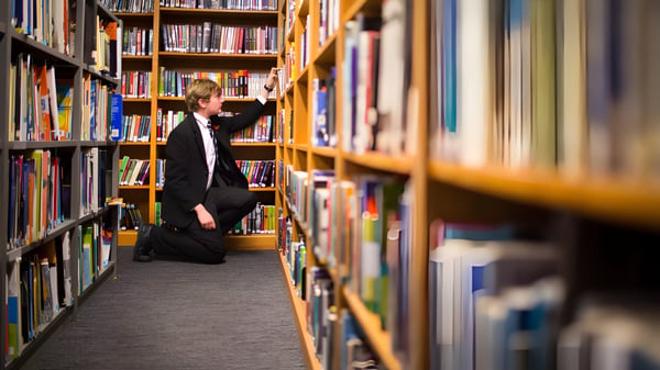Eine Person im schwarzen Mantel steht im engen Gang zwischen hohen Bücherregalen in der Bibliothek vom Campbell College.