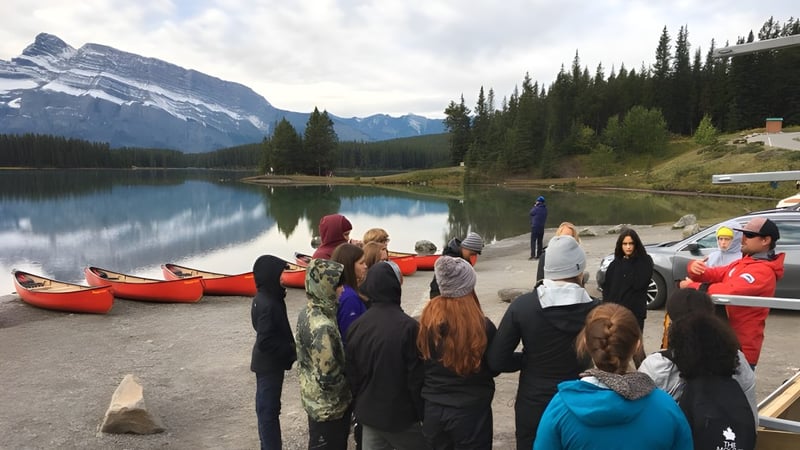 Eine Gruppe von Schülern am Ufer eines Sees mit Bergen und Wald im Hintergrund auf dem Gelände der Canmore Collegiate High School.