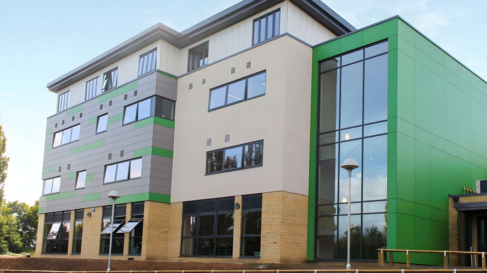 Das moderne mehrstöckige Gebäude der Canterbury Academy mit beige-grünem Äußeren und großen Fenstern vor klarem blauen Himmel.