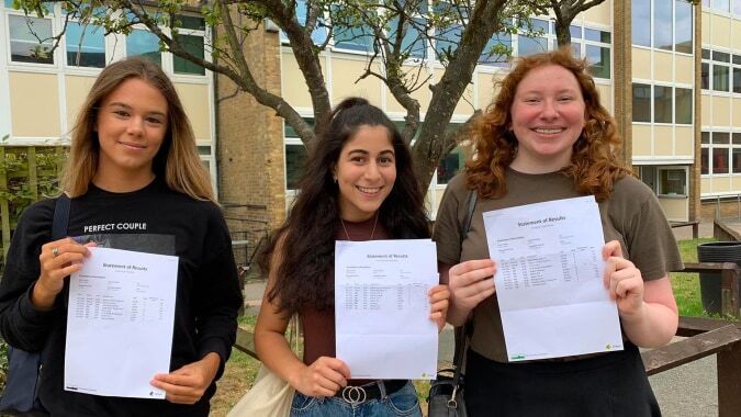 Drei Schülerinnen der Canterbury Academy halten gemeinsam ihre akademischen Dokumente vor einem Schulgebäude mit Bäumen.