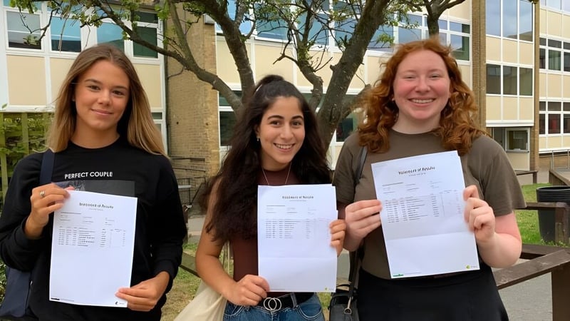 Drei Schülerinnen der Canterbury Academy zeigen im Freien ihre akademischen Unterlagen vor dem Schulgebäude.