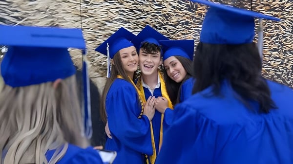 Absolventen der Canterbury High School feiern in blauen Talaren und Hüten ihren Abschluss vor einem Hintergrund mit Bäumen.
