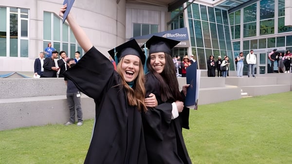 Zwei Absolventinnen der Capilano University umarmen sich vor einem Universitätsgebäude mit weiteren Graduierenden im Hintergrund.