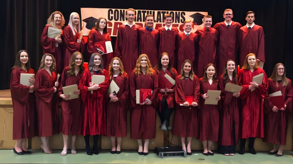 Eine Gruppe von Absolventen der Carbonear Collegiate High School steht in roten Talaren auf einer Bühne mit einem Banner mit der Aufschrift ‚Congratulations‘.
