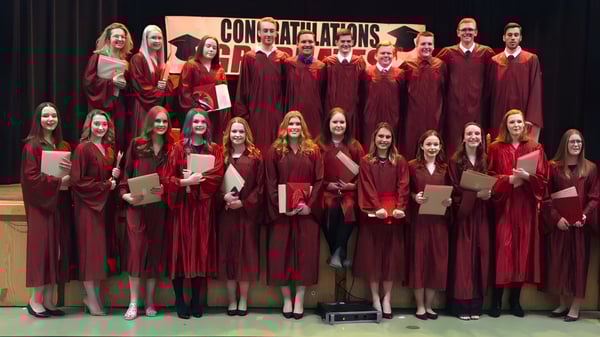 Eine Gruppe von Absolventen der Carbonear Collegiate High School steht in roten Talaren auf einer Bühne mit einem Banner mit der Aufschrift ‚Congratulations‘.