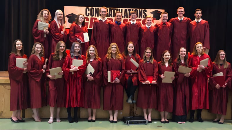 Eine Gruppe von Absolventen der Carbonear Collegiate High School steht in roten Talaren auf einer Bühne mit einem Banner mit der Aufschrift ‚Congratulations‘.