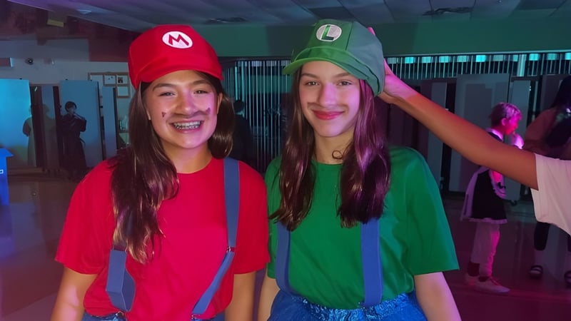 Zwei Schülerinnen der Carrefour de l’Acadie tragen bunte Kostüme und posieren bei einer Faschingsfeier im Schulgebäude.