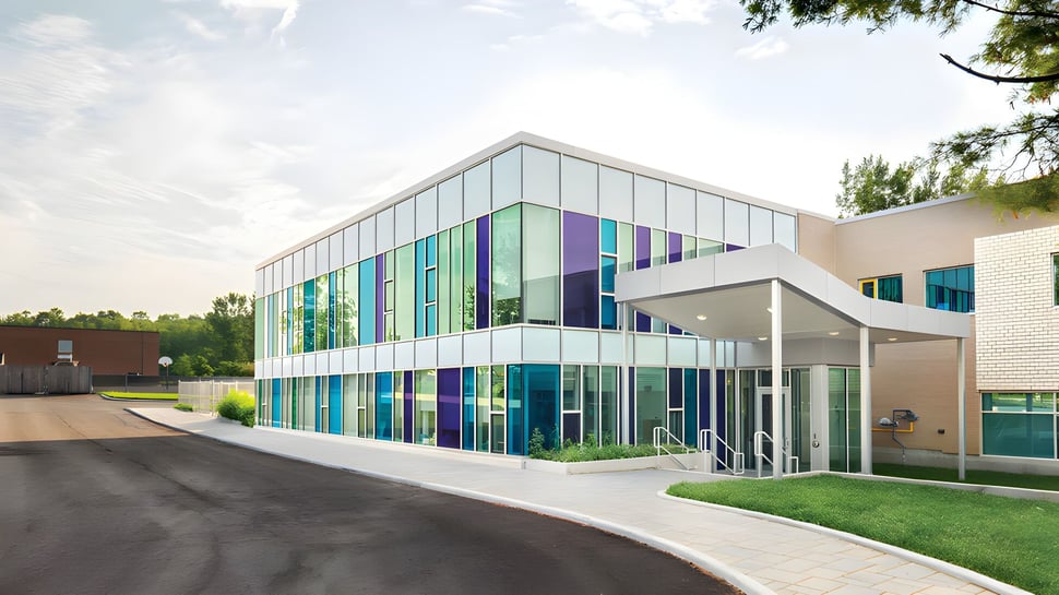 Das mehrstöckige Schulgebäude der École secondaire publique Carrefour Jeunesse mit Glasfassade und farbigen Paneelen steht in einer begrünten Umgebung.