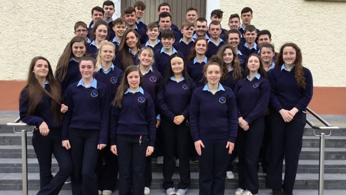 Eine Gruppe von Schülerinnen und Schülern der Carrigallen Vocational School steht mit blauen Schuluniformen vor einem Gebäude mit Backsteinmauer.