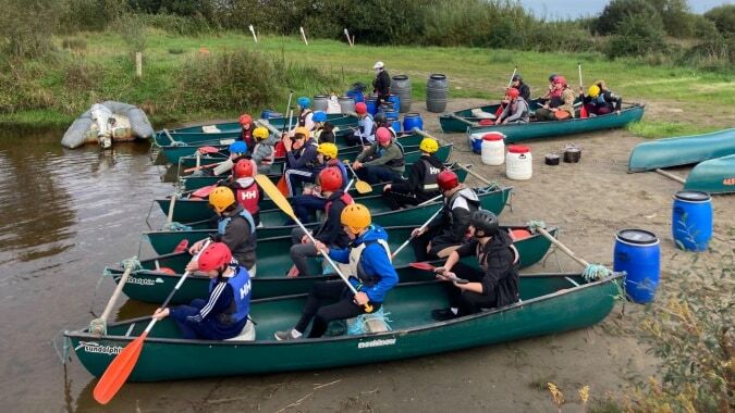 Schüler der Carrigallen Vocational School sitzen in grünen Kanus auf einem Fluss umgeben von üppiger Vegetation.