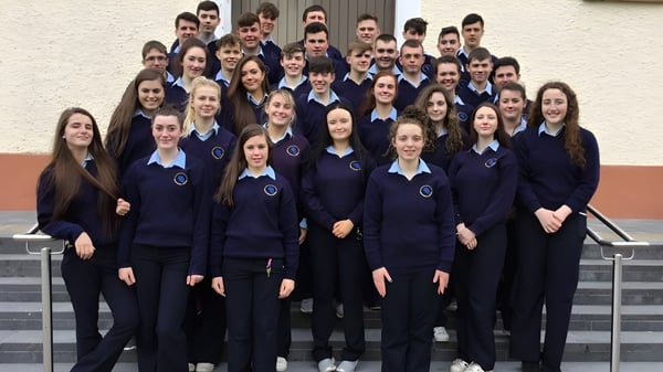 Eine Gruppe von Schülerinnen und Schülern in einheitlichen dunkelblauen Uniformen steht vor einer weißen Wand auf dem Gelände der Carrigallen Vocational School.