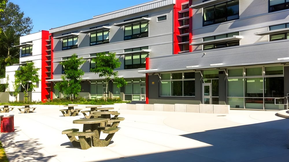 Das mehrstöckige moderne Gebäude der Carson Graham Secondary School mit großen Fenstern und roten Akzenten steht hinter einem gepflasterten Hof mit Holzbänken.