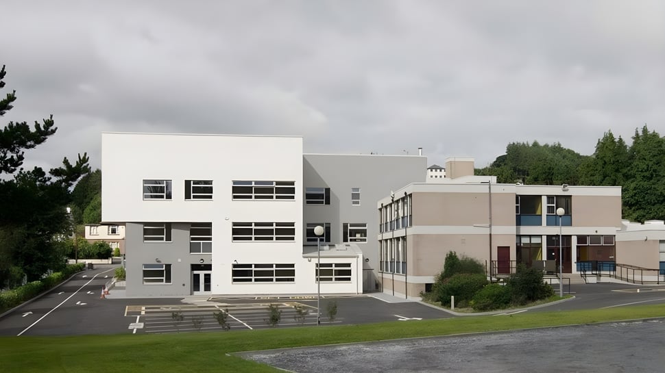 Das moderne mehrstöckige Gebäude von Castleblayney College steht umgeben von Rasen und Bäumen unter bewölktem Himmel.