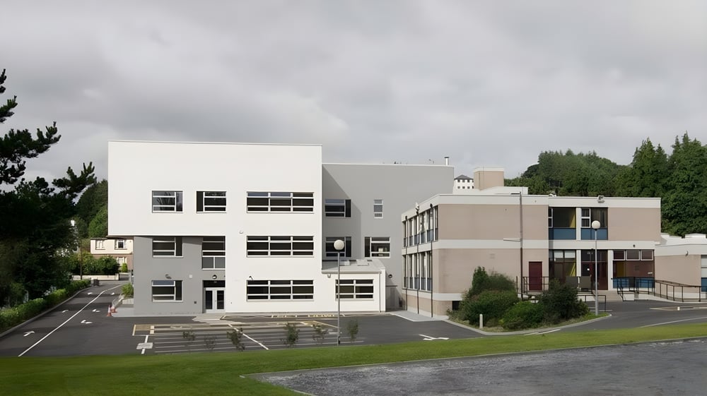 Das moderne mehrstöckige Gebäude von Castleblayney College steht umgeben von Rasen und Bäumen unter bewölktem Himmel.