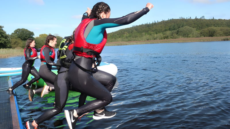 Eine Gruppe von Schülerinnen und Schülern des Castleblayney College steht auf einem Paddleboard auf einem See, umgeben von bewaldeten Hügeln.