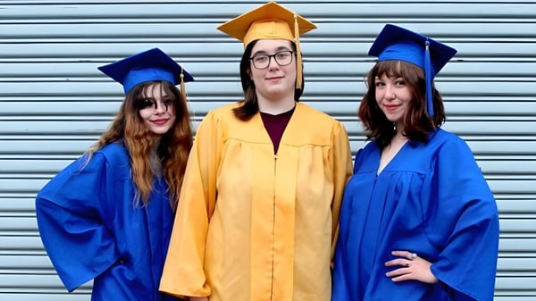 Drei Personen in verschiedenen Abschlussroben stehen vor einem Metallrollladen auf dem Campus des Castlepollard Community College.