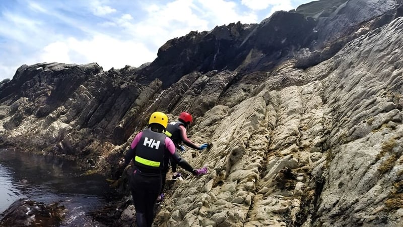 Eine Schülerin oder ein Schüler der Castlerea Community School klettert mit einem gelben Helm an einer felsigen Klippe mit Blick auf Wasser und Berge.
