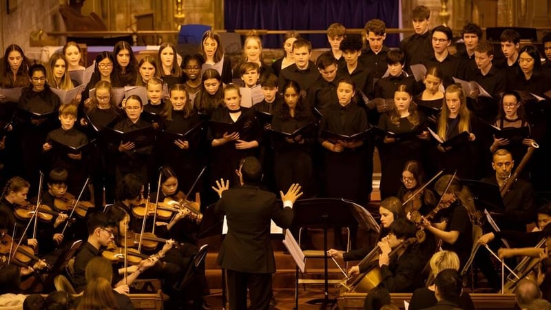 Ein großer Chor in schwarzen Roben singt auf der Bühne mit einem Orchester im Vordergrund bei Caterham School.