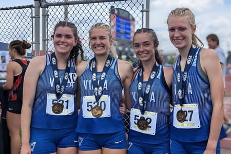 Vier Schülerinnen der Cathedral Preparatory School stehen in Sportuniformen neben dem eingezäunten Leichtathletikstadion.